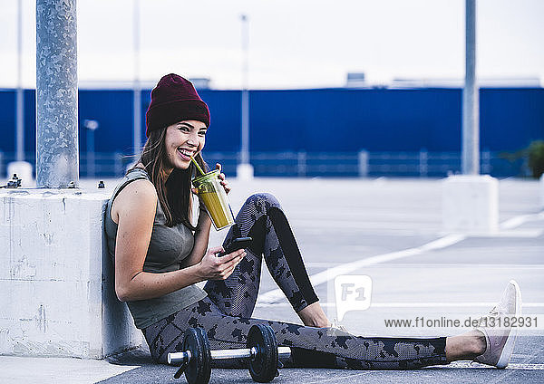 Junge Frau sitzt nach Hanteltraining am Boden und benutzt ein Smartphone