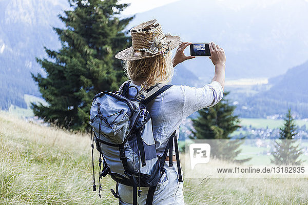 Deutschland  Bayern  Oberammergau  junge Frau wandert und macht ein Handyfoto auf einer Bergwiese