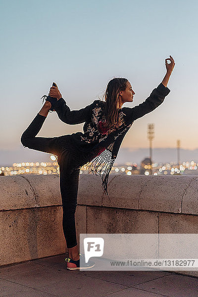 Spain  Barcelona  Montjuic  young woman doing yoga at dusk with city lights in background