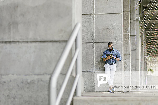 Bearded hipster businessman leaning against concrete wall using tablet