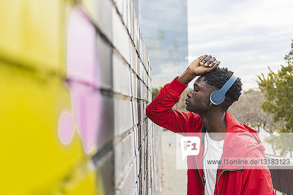 Junger schwarzer Mann lehnt an der Wand und hört Musik mit Kopfhörern