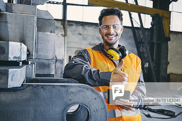 Porträt eines lächelnden Mannes in Arbeitsschutzkleidung mit Daumen nach oben in der Fabrik
