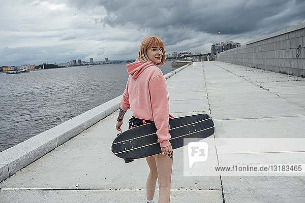 Junge Frau mit Skateboard in der Hand beim Gehen am Flussufer