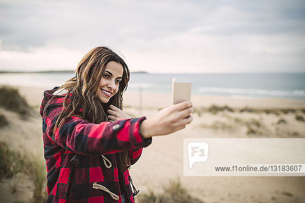 Porträt einer lächelnden jungen Frau  die sich am Strand mit einem Smartphone selbstständig macht