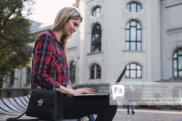 Modische junge Frau  die im Freien auf einer Bank sitzt und am Laptop arbeitet