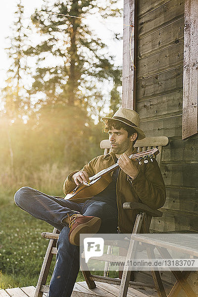Junger Mann sitzt auf der Veranda eines Holzhauses und spielt Ukulele
