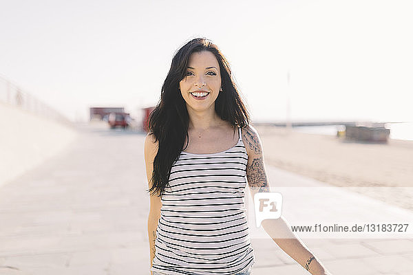 Porträt einer lächelnden jungen Frau mit Tätowierung beim Spaziergang an der Strandpromenade
