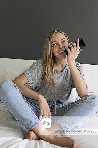 Glückliche junge Frau sitzt auf dem Bett und benutzt ihr Handy