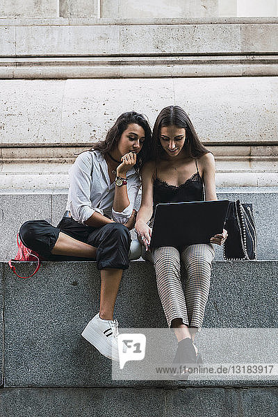 Two friends sitting side by side on a wall using laptop