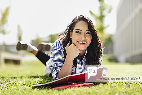 Porträt eines lächelnden Studenten  der mit Notizbüchern auf dem Rasen liegt