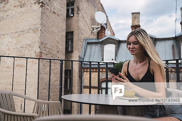 Lächelnde blonde Frau sitzt mit einer Tasse Kaffee auf der Dachterrasse und überprüft Handy
