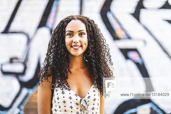 Portrait of smiling young woman with long curly hair at graffiti wall
