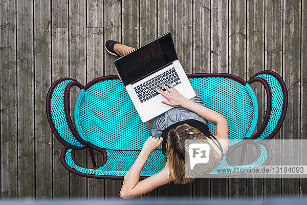 Junge Frau sitzt auf türkisfarbener Couch auf der Terrasse mit Laptop  Draufsicht
