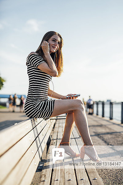 USA  New York  Brooklyn  Frau sitzt auf einer Bank und hört Musik mit Smartphone und Kopfhörern