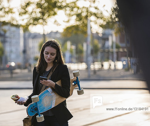 Young woman with longboard and snack in the city checking cell phone