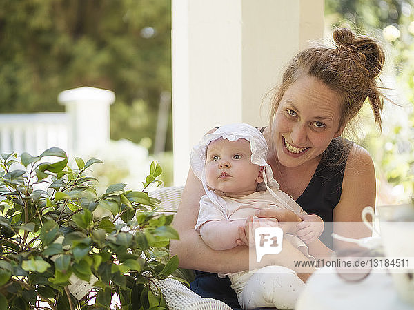Porträt einer lächelnden Mutter mit einem auf dem Schoß sitzenden Mädchen auf der Terrasse