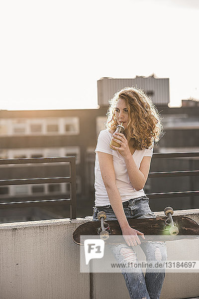 Junge Frau mit Skateboard trinkt Getränk auf der Dachterrasse bei Sonnenuntergang