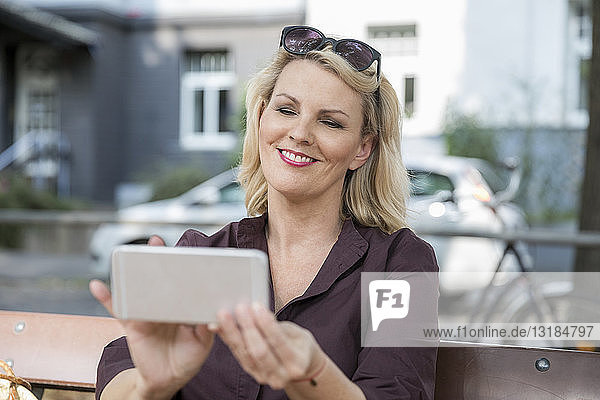 Porträt einer lächelnden  blonden  reifen Frau  die ein Smartphone im Freien benutzt