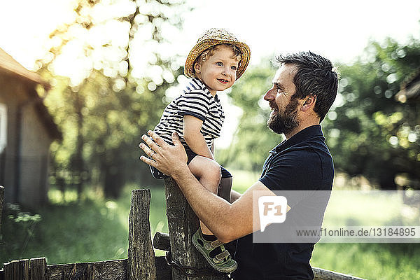 Glücklicher Vater schaut auf seinen lächelnden kleinen Sohn  der auf einem Zaun sitzt
