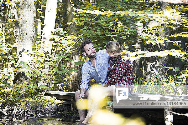 Junges Paar sitzt auf einer Brücke im Wald mit Füßen im Wasser