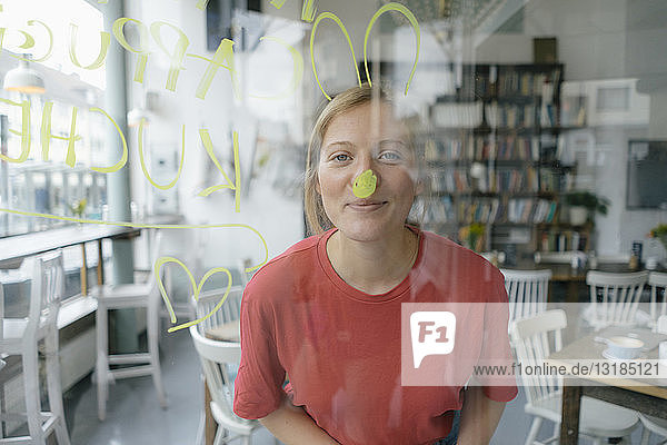 Porträt einer lächelnden jungen Frau  die hinter einer Fensterscheibe in einem Cafe posiert