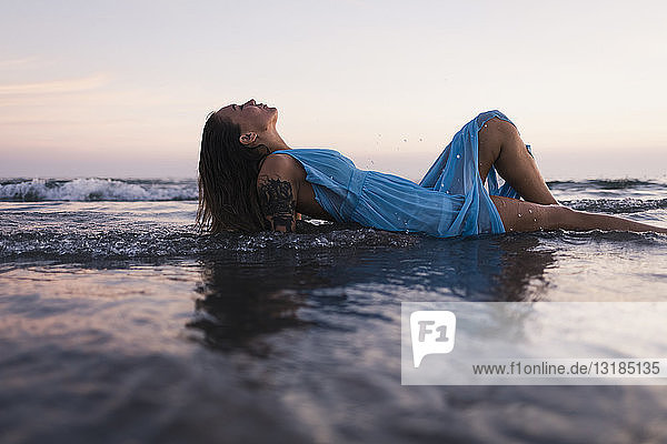Junge tätowierte Frau in blauem Kleid liegt bei Sonnenuntergang im Wasser am Meeresufer