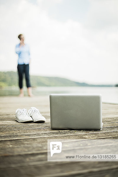 Laptop und Schuhe auf einem Steg an einem See mit einer Frau im Hintergrund
