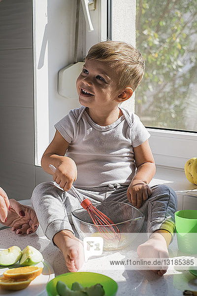 Porträt eines lächelnden kleinen Jungen  der barfuss auf der Arbeitsplatte in der Küche sitzt und Eier verquirlt