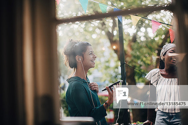 Glückliche junge Frau spricht durch Kopfhörer  während sie während einer Party mit einem Freund auf dem Balkon steht