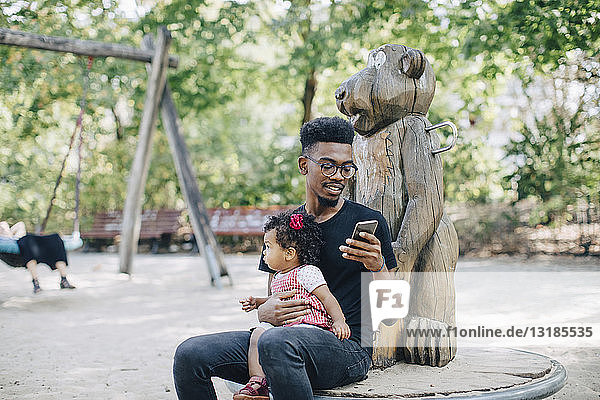 Vater benutzt Mobiltelefon  während er mit der Tochter auf Spielgeräten im Freien sitzt