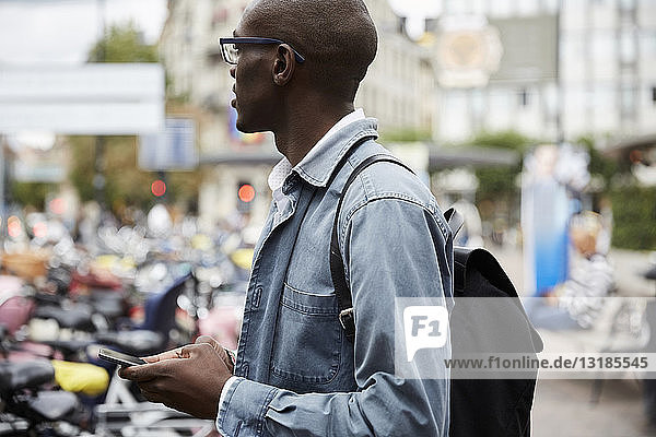 Seitenansicht eines mittelgroßen  erwachsenen Geschäftsmannes  der weg schaut  während er mit einem Smartphone in der Stadt steht