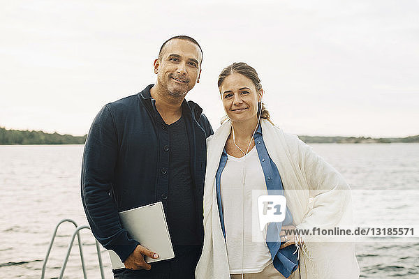 Portrait of smiling mature couple with laptop standing against lake