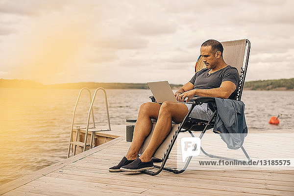 Erwachsener Mann in voller Länge mit Laptop  während er auf einem Liegestuhl am Steg sitzt