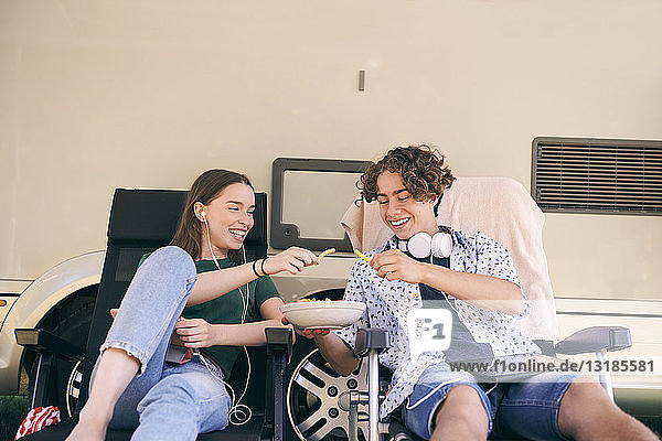 Fröhliche Geschwister genießen Snacks  während sie auf Stühlen gegen einen Lieferwagen auf dem Campingplatz sitzen