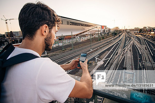Young man using mobile phone while leaning on railing over railroad tracks in city