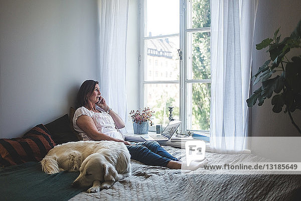 Dog sleeping and woman talking on phone while looking through window in bedroom at home