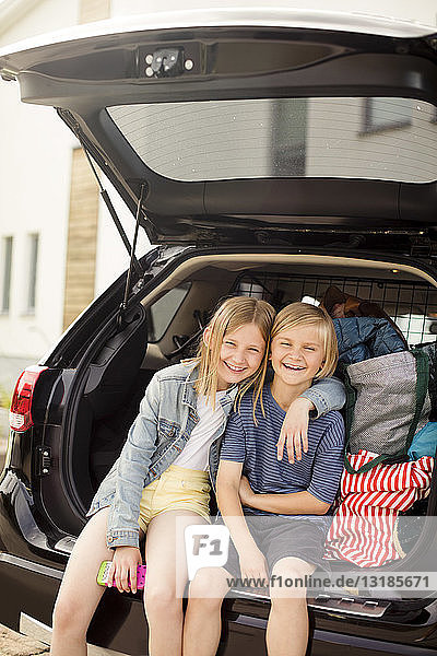 Porträt von lächelnden Geschwistern  die im Autokofferraum vor dem Haus sitzen