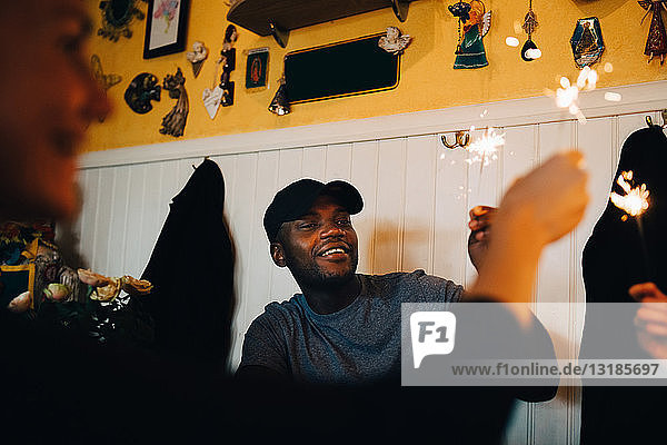 Lächelnder junger Mann mit brennender Wunderkerze in der Hand  während ein Freund während der Dinnerparty im Restaurant sitzt