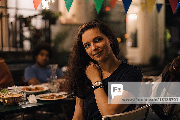 Porträt einer lächelnden jungen Frau  die während einer Dinnerparty im Hinterhof auf einem Stuhl am Tisch sitzt
