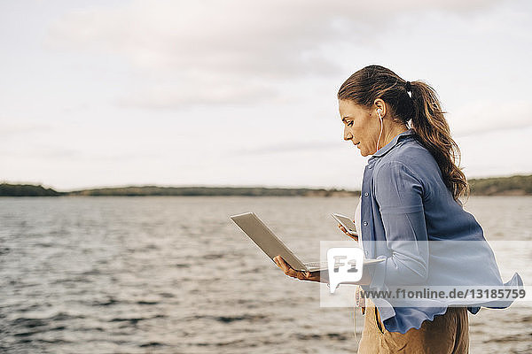 Selbstbewusste  reife Frau  die einen Laptop benutzt  während sie am See mit dem Handy telefoniert