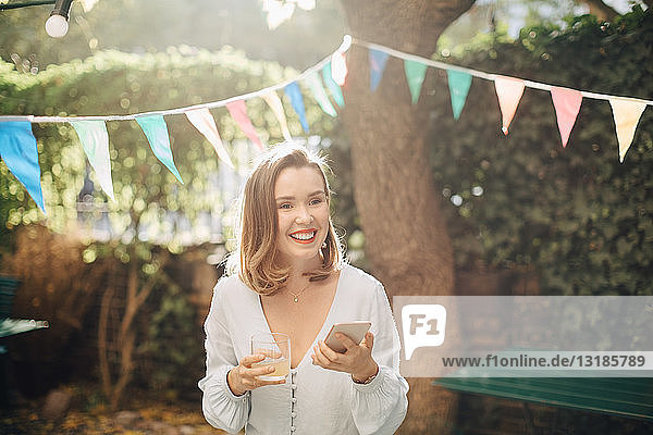 Smiling young woman holding drink and mobile phone while standing in backyard