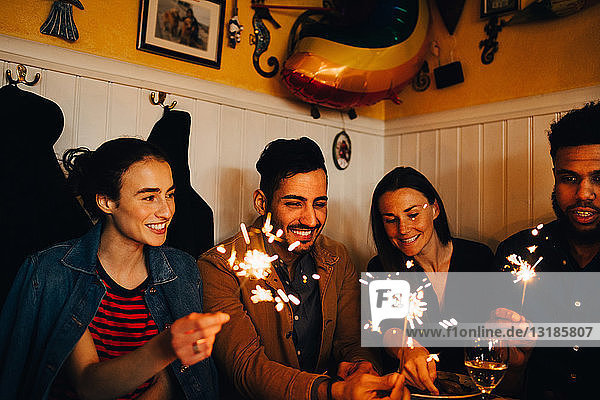 Glückliche junge multiethnische Freunde und Freundinnen mit brennenden Wunderkerzen im Restaurant während der Dinnerparty