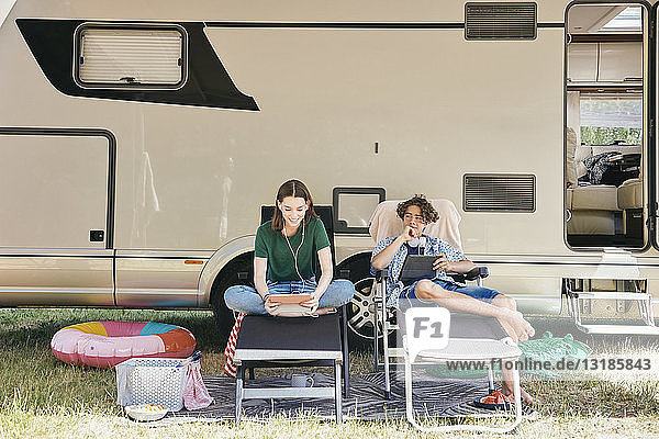 Bruder und Schwester benutzen ein digitales Tablett  während sie auf Klappstühlen gegen ein Wohnmobil sitzen