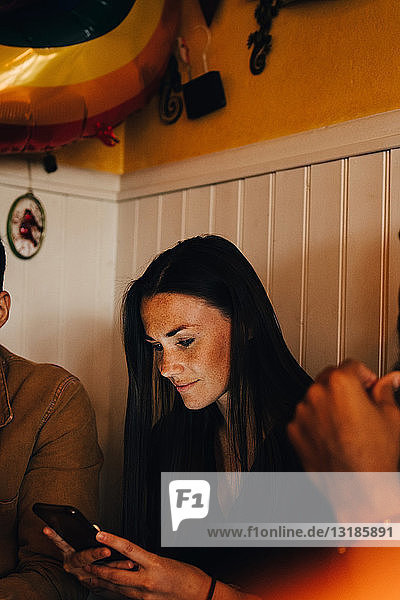 Junge Frau benutzt Smartphone  während sie inmitten multiethnischer männlicher Freunde im Restaurant sitzt