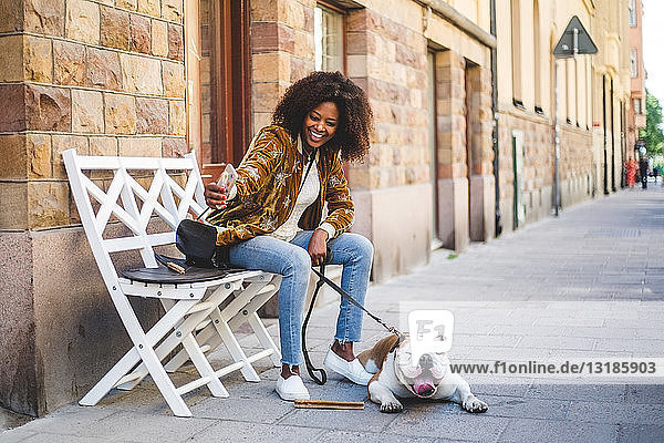 Glückliche Frau sitzt mit Hund  der sich auf dem Bürgersteig in der Stadt mit seinem Handy selbstständig macht