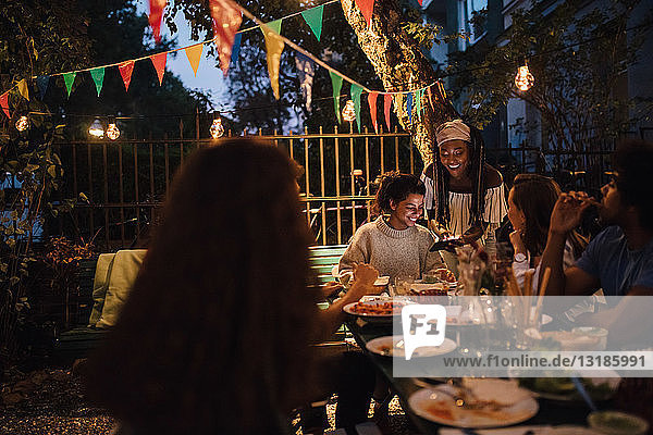 Lächelnde junge Frau zeigt Freundinnen bei Tisch während einer Dinnerparty ihr Handy