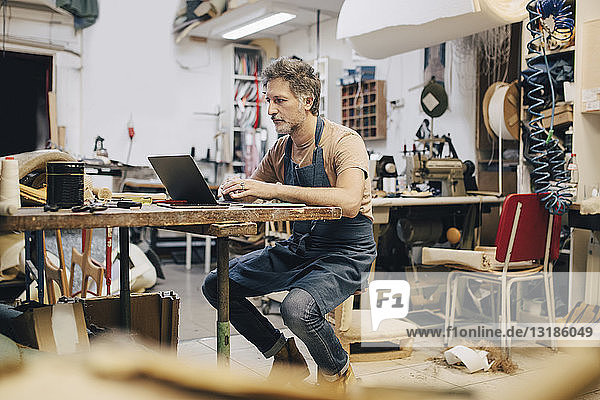 Male upholstery worker using laptop at workbench in workshop