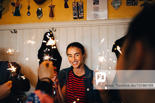 Lächelnde junge multiethnische Freunde halten brennende Wunderkerzen in der Hand  während sie während der Dinnerparty im Restaurant genießen