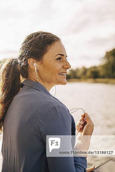 Lächelnde Frau  die am See sitzend über Kopfhörer mit einem digitalen Tablet spricht