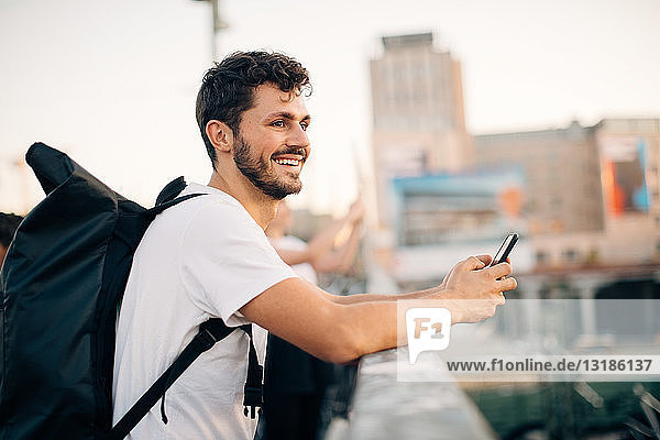 Seitenansicht eines lächelnden jungen Mannes  der wegschaut  während er sein Mobiltelefon an der Brücke hält